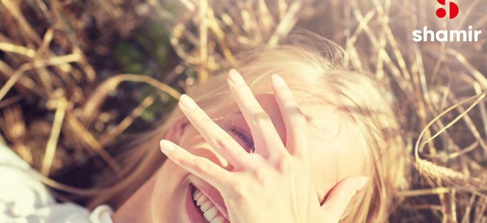 Mujer sonriente al aire libre cubriéndose el rostro parcialmente con la mano, mientras disfruta de la luz del sol en un entorno natural. Imagen representativa de la tecnología Shamir Transitions®, que adapta automáticamente las lentes a los cambios de luz para proteger la vista. Logo de Shamir visible en la esquina superior derecha.