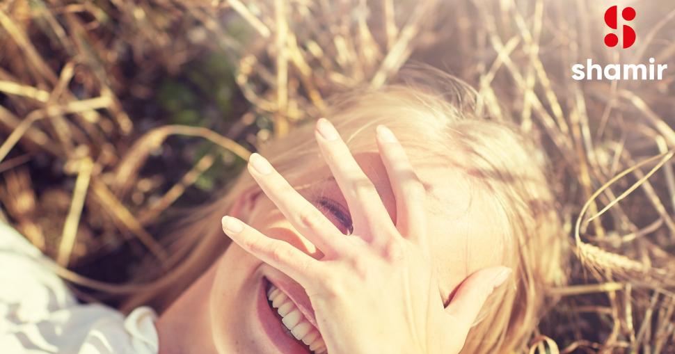 Mujer sonriente al aire libre cubriéndose el rostro parcialmente con la mano, mientras disfruta de la luz del sol en un entorno natural. Imagen representativa de la tecnología Shamir Transitions®, que adapta automáticamente las lentes a los cambios de luz para proteger la vista. Logo de Shamir visible en la esquina superior derecha.