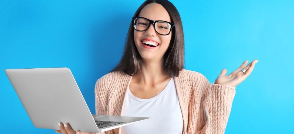 Mujer sonriente con gafas y un ordenador portátil frente a un fondo azul, demostrando estilo y protección ocular mientras trabaja