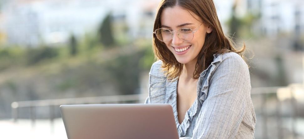 Mujer joven con gafas trabajando en su portátil al aire libre, disfrutando de una vista relajante mientras protege sus ojos de la luz azul.