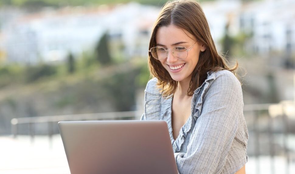 Mujer joven con gafas trabajando en su portátil al aire libre, disfrutando de una vista relajante mientras protege sus ojos de la luz azul.
