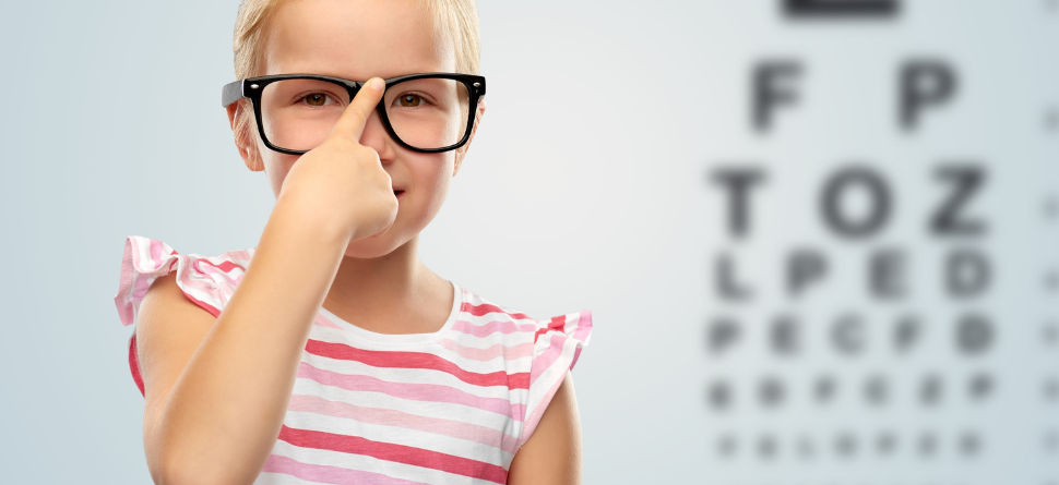 Niña pequeña con gafas señalando su nariz frente a un gráfico de visión, resaltando la importancia de las revisiones oculares en niños.