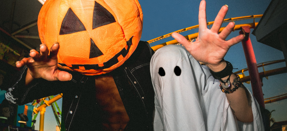 Dos personas disfrazadas en una feria, una con cabeza de calabaza y otra como fantasma, posando de forma divertida. Las gafas pueden ser un accesorio perfecto para completar estos disfraces al aire libre y proteger los ojos de luces intensas o efectos especiales.