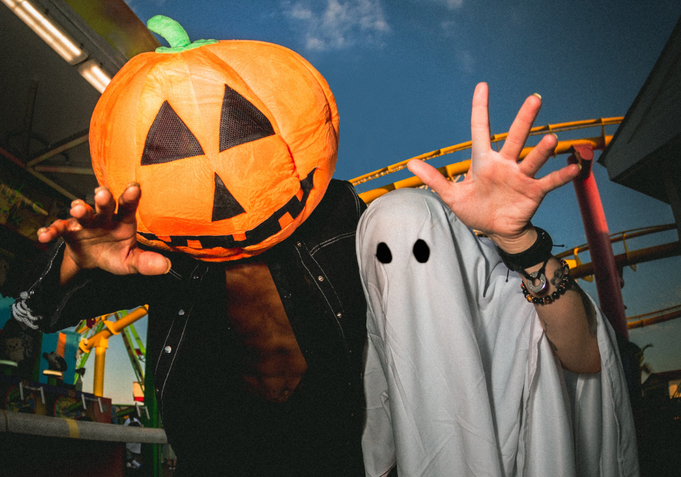 Dos personas disfrazadas en una feria, una con cabeza de calabaza y otra como fantasma, posando de forma divertida. Las gafas pueden ser un accesorio perfecto para completar estos disfraces al aire libre y proteger los ojos de luces intensas o efectos especiales.