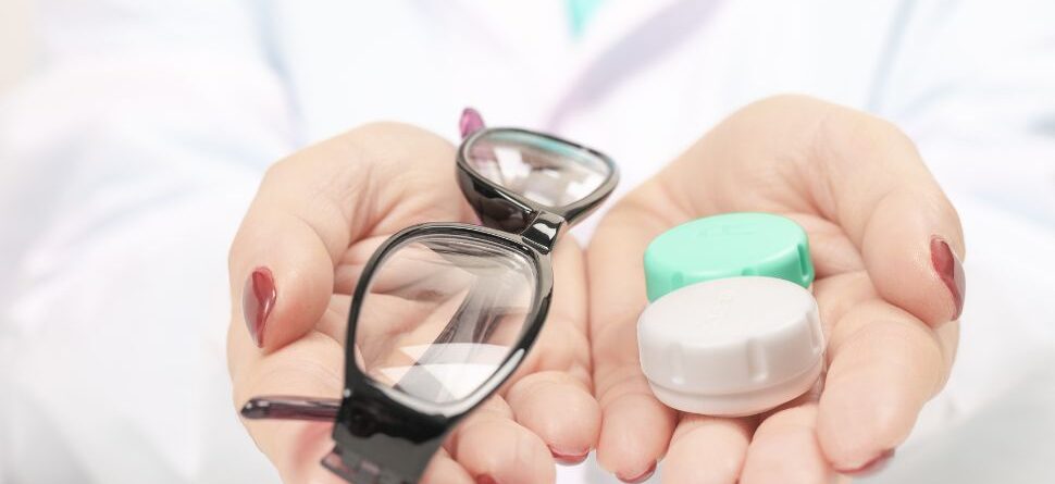 Primer plano de una persona con bata blanca que sostiene en la mano izquierda unas gafas de montura negra y en la mano derecha un estuche de lentillas. La imagen representa la comparación entre gafas y lentillas para el cuidado visual.