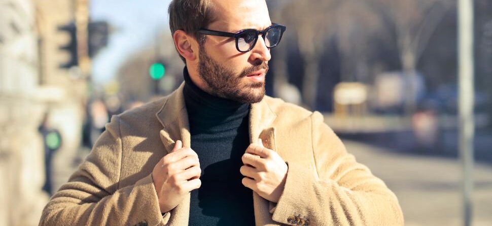 Hombre con barba y gafas de sol oscuras caminando por una ciudad, vistiendo un abrigo beige y jersey negro. La imagen muestra la elegancia y protección de las gafas de sol en exteriores.