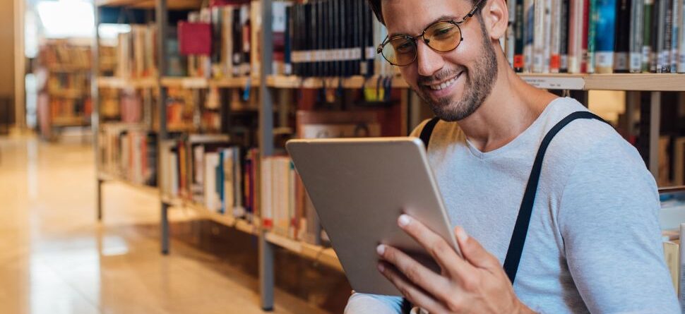 Imagen de un hombre con gafas en una biblioteca leyendo un dispositivo: Un hombre sonriente con gafas de lectura modernas está sentado en una biblioteca mientras lee en su tablet. La imagen resalta la practicidad y el diseño de las gafas de lectura, ideales para quienes combinan lectura física y digital.