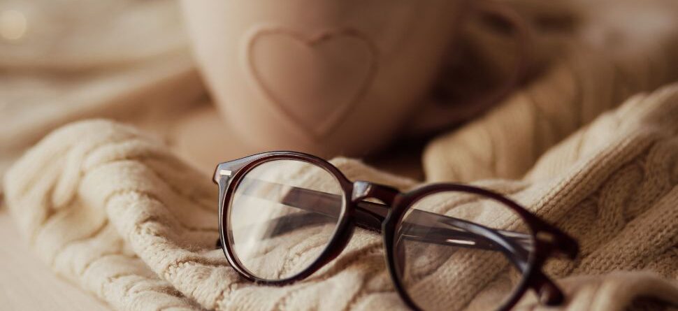 Imagen de gafas sobre un jersey y una taza de café: Un par de gafas de lectura descansa sobre un jersey tejido junto a una taza de café, en un ambiente cálido con luces navideñas desenfocadas de fondo. Transmite confort y la conexión entre lectura y momentos de relajación durante el invierno.
