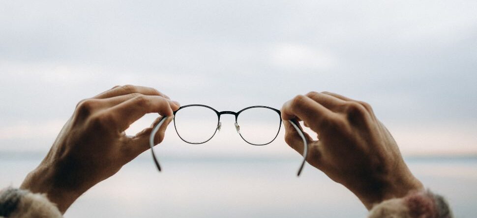 Primer plano de unas gafas sostenidas por dos manos frente a un paisaje difuminado, simbolizando claridad visual y enfoque.