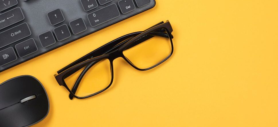 Gafas negras colocadas sobre un escritorio amarillo junto a un teclado y un ratón, representando el cuidado visual en entornos laborales digitales.