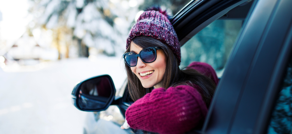 Mujer con gafas de sol asomada por la ventanilla de un coche en un entorno invernal. La protección UV es fundamental en cualquier clima.