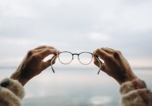 Primer plano de unas gafas sostenidas con ambas manos frente a un fondo desenfocado, representando la claridad visual que aportan unas gafas de calidad óptica en Óptica Cosmos Tenerife.