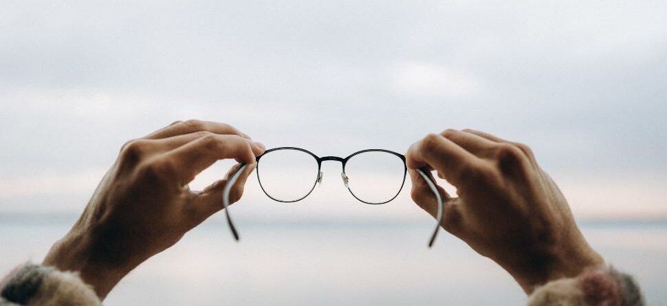 Primer plano de unas gafas sostenidas con ambas manos frente a un fondo desenfocado, representando la claridad visual que aportan unas gafas de calidad óptica en Óptica Cosmos Tenerife.