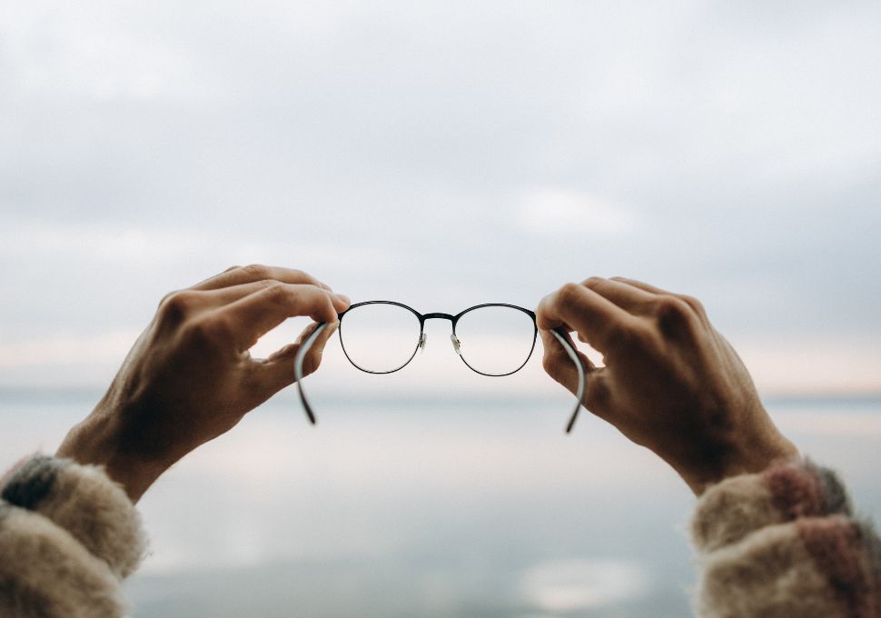 Primer plano de unas gafas sostenidas con ambas manos frente a un fondo desenfocado, representando la claridad visual que aportan unas gafas de calidad óptica en Óptica Cosmos Tenerife.