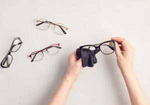 Persona limpiando gafas con un paño de microfibra junto a varios modelos de gafas sobre fondo claro, destacando el mantenimiento de gafas en Óptica Cosmos Tenerife.
