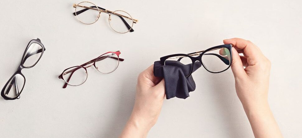 Persona limpiando gafas con un paño de microfibra junto a varios modelos de gafas sobre fondo claro, destacando el mantenimiento de gafas en Óptica Cosmos Tenerife.